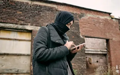 Osoba w czarnej chuście i kurtce trzyma telefon na tle zrujnowanej ceglanej ściany.