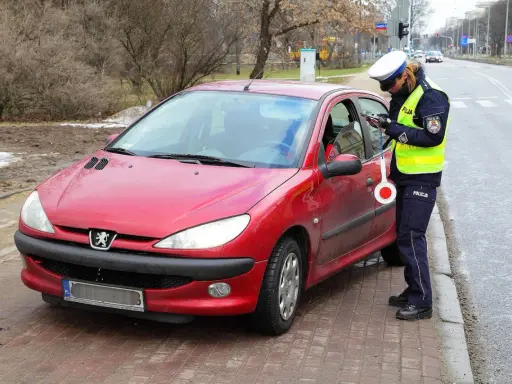 Policjantka w kamizelce odblaskowej i czapce spisuje kierowcę przy czerwonym samochodzie.