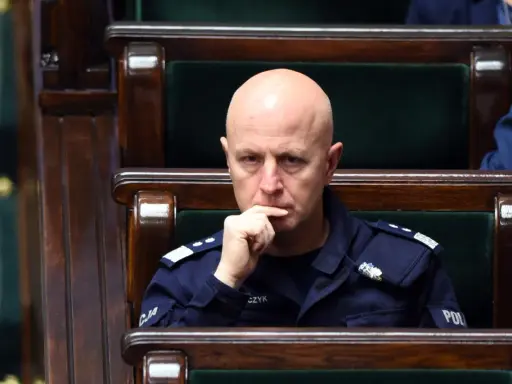 Łysy mężczyzna w mundurze policji siedzi w drewnianej ławie, zamyślony.
