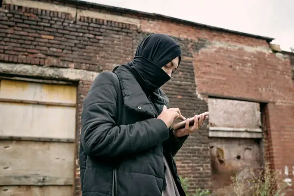 Osoba w czarnej chuście i kurtce trzyma telefon na tle zrujnowanej ceglanej ściany.